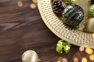Close-up golden round tray with green and gold Christmas tree balls and pine cones on wooden background. Christmas background with copy space. Gerland defocused lights. Preparation for the New Year.