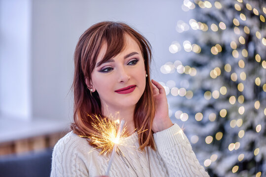 Portrait of a young woman in a white knitted long sweater dress, holding Bengal light in hands. Blurred background of blue Christmas tree with bokeh lights from garlands. New Year's festive interior
