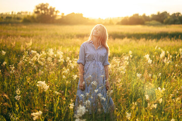 a beautiful woman stands in a field at sunset in the light of the sun in a beautiful blue dress
