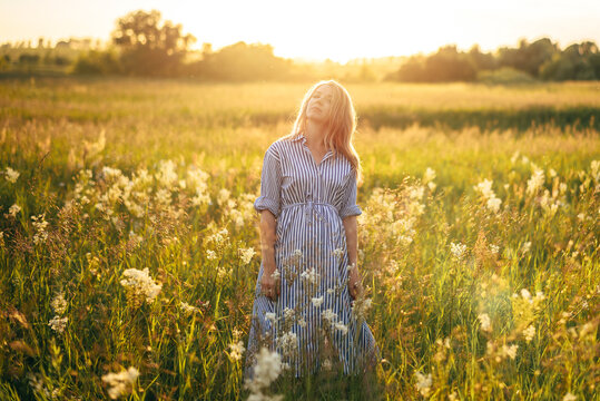 A Beautiful Woman Stands In A Field At Sunset In The Light Of The Sun In A Beautiful Blue Dress
