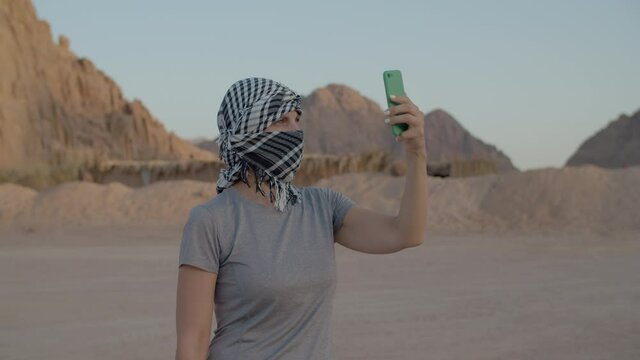 Woman in checkered keffiyeh trying to find mobile network using mobile phone standing in desert. Person searching for mobile signal to make a call in desert. 