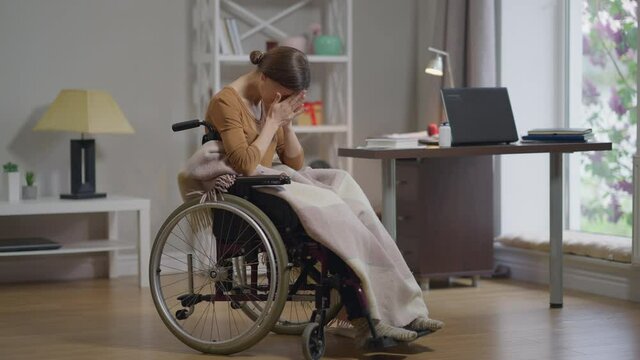 Overwhelmed young disabled woman in wheelchair closing face crying alone at home. Wide shot portrait of depressed sad Caucasian lady holding head in hands. Hopelessness and frustration concept