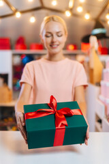 Vertical shot of smiling attractive young woman offering take Christmas gift box tied red ribbon and decorated beautiful bow standing at counter of holiday store, background of festive packages.
