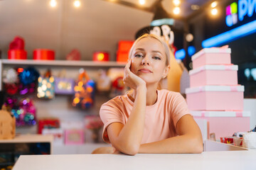 Front view of frustrated young sales woman sitting at counter of holiday store in mall waiting for...
