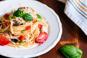 Fresh pasta with spaghetti, tomato, mushrooms, basil and parmesan cheese in white plate on a wooden background with ingredients in background