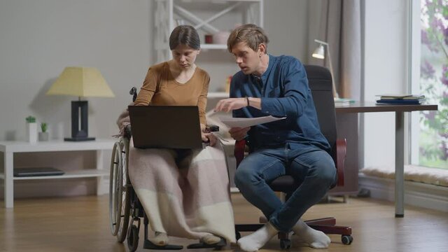 Front view concentrated serious Caucasian woman in wheelchair and man talking analyzing paperwork surfing Internet on laptop. Young couple of freelancers working at home indoors in slow motion
