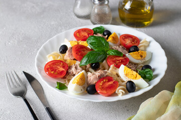 Canned tuna salad with pasta, cherry tomatoes, black olives, boiled egg and basil on gray background. Healthy food.