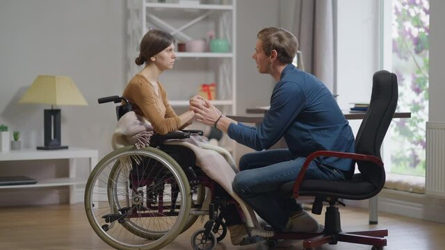 Side view confident loving man supporting depressed sad woman in wheelchair. Wide shot Caucasian husband talking to desperate young wife in slow motion holding hands. Family unity and endorsement