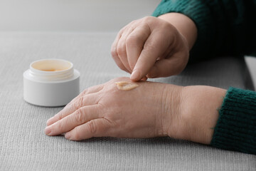Fototapeta na wymiar Elderly woman applying cosmetic cream onto her hands at table