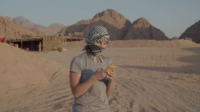 Female tourist in checkered keffiyeh standing in the desert using mobile phone. Caucasian woman taking off headwear and smiling at camera in desert.