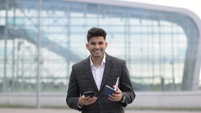 Arab businessman waiting flight standing outside airport. Handsome hindu man holding cell phone, passport and boarding pass checking his flight schedule.