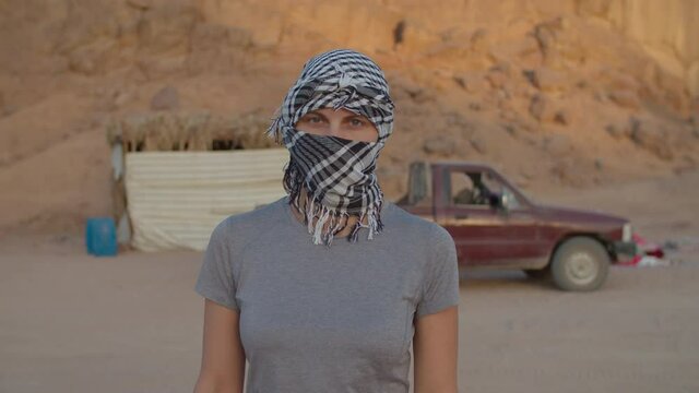 Portrait of woman wearing black and white checkered keffiyeh standing in the desert looking at camera. Caucasian female tourist taking off headdress and smiling at camera.