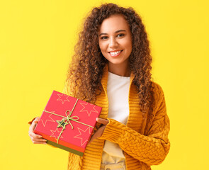 Young African-American woman in warm sweater with Christmas present on yellow background