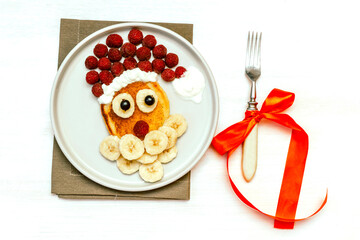 Christmas Santa Claus shaped pancake with sweet fresh raspberry berry and banana on plate on white wooden background for kids children breakfast. xmas food with new year decorations with copy space