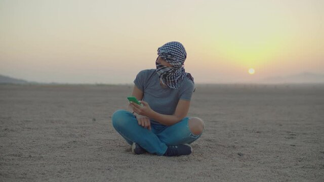 Woman in checkered keffiyeh sitting on desert sand using mobile phone. Caucasian female tourist enjoy desert adventure during sunrise.