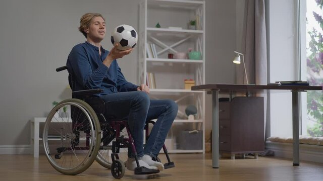 Thoughtful smiling disabled young man in wheelchair with soccer ball indoors at home. Wide shot portrait confident motivated injured Caucasian sportsman thinking on recovery. Slow motion