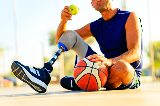 sporty handicapped man in sportswear and with artificial leg basketball player at sunset eating an apple