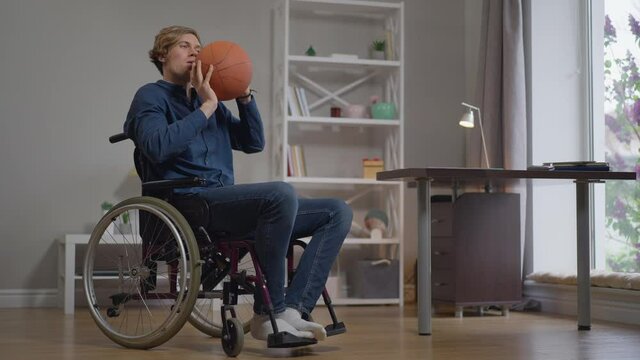 Young sportsman in wheelchair throwing basketball ball in slow motion training at home indoors. Portrait of inspired Caucasian disabled man exercising dreaming of paralympic games