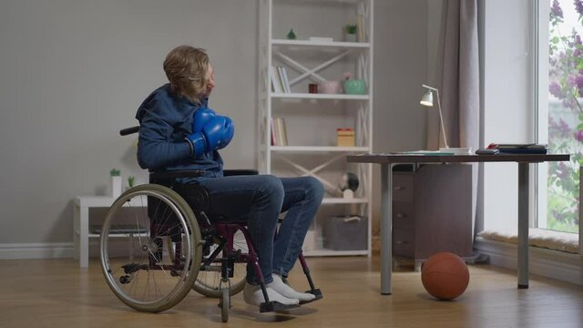 Young depressed boxer in wheelchair training taking off boxing gloves holding head in hands. Frustrated Caucasian disabled man at home indoors alone. Sport and disability