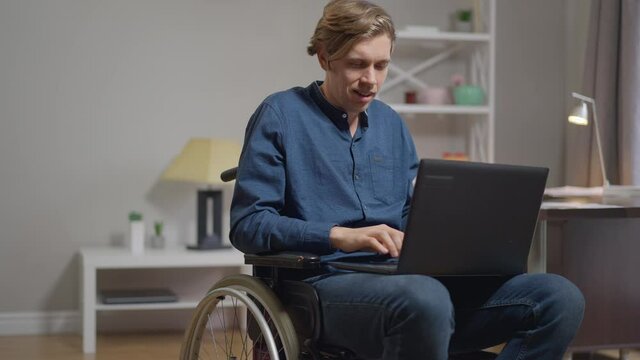 Portrait of confident absorbed young disabled man texting on laptop keyboard looking at camera smiling. Satisfied happy Caucasian freelancer in wheelchair posing indoors at home in slow motion