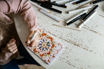 A little cute girl draws a circular mandala pattern in the album with art markers.