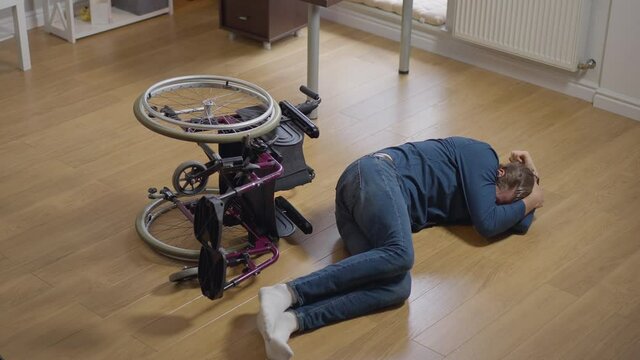 High angle wide shot desperate crying disabled man lying on floor with wheelchair rolled over. Young devastated Caucasian paralyzed helpless guy indoors at home alone. Slow motion
