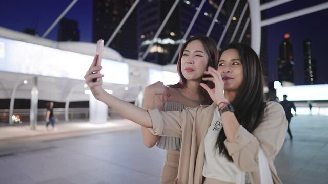 Two attractive asian female tourist making video calling to other friend showing a beautiful modern cityscape behind, distant communication, wireless internet connection and people, holiday vacation 