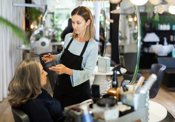 Fototapeta na wymiar Skillful woman hair stylist making haircut to aged female client in hairdressing studio.