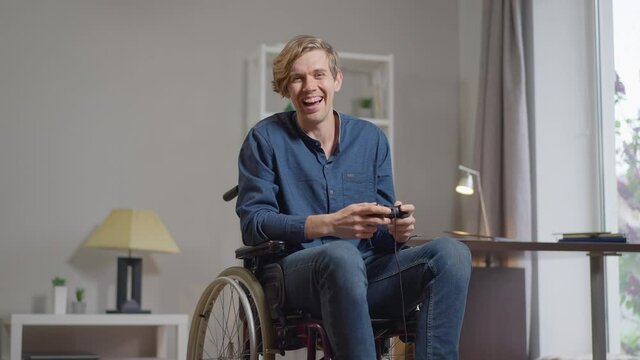 Portrait of joyful Caucasian young handicapped man in wheelchair winning in multiplayer online video game gesturing victory in slow motion. Happy disabled gamer with joystick rejoicing indoors