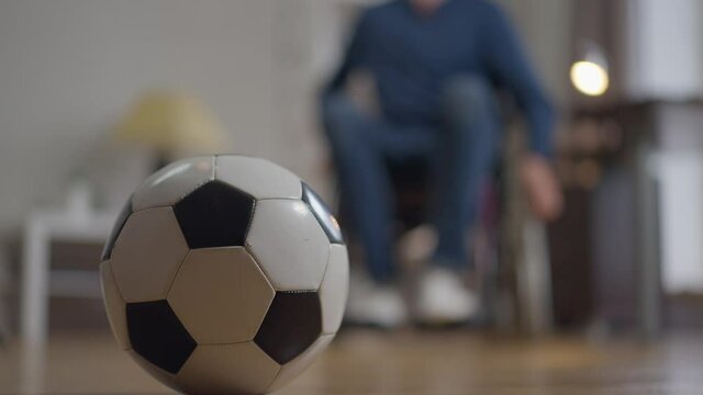 Close-up soccer ball with disabled blurred man rolling wheelchair from background pushing sport equipment. Unrecognizable paraplegic Caucasian sportsman at home indoors. Paralympic concept