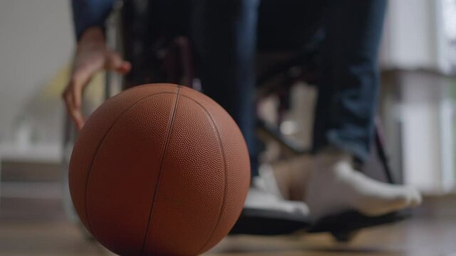 Basketball ball in living room indoors with unrecognizable blurred disabled young man rolling wheelchair leaving with ball. Paraplegic paralympic sportsman indoors. Slow motion