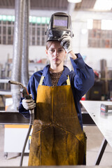 Young workwoman skilled welder wearing protective apron, gloves and helmet standing in metalworking...