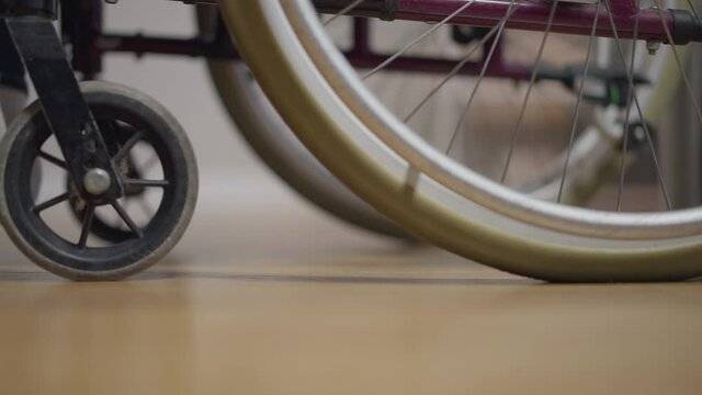 Side view wheelchair with disabled man and woman pushing in slow motion. Unrecognizable paraplegic guy with caregiver indoors at home or in nursing home