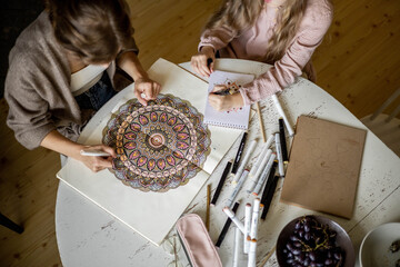 Young beautiful mother and cute daughter draw a mandala pattern together. Artistic markers and colored pencils.