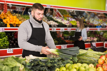 Male shop assistant in apron lays fresh leek in supermarket. High quality photo