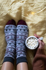 A large mug of cocoa in the hands of a girl in a warm sweater, sitting in warm socks on the bed. Top view.