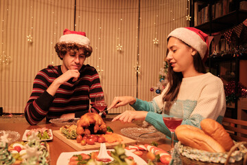 A young couple enjoys eating at table with specials foods, girlfriend cutting roasted turkey at home's dining room, decorated with ornaments, Christmas festival, and New Year celebration party.