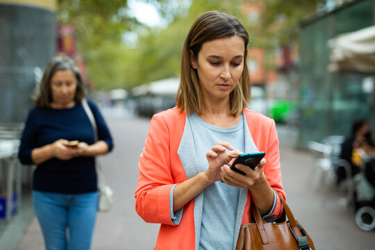 Focused young business woman using smartphone to deal with work issues while walking along city street in warm autumn day .