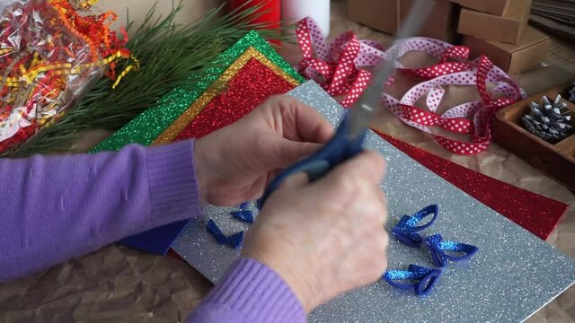 The Woman's Hands Cut Out The Snowflake Element From Blue Glitter Foamiran, On The Table Colorful Foamiran Sheets, Red And White Ribbon, Cones. Preparing For Christmas. DIY