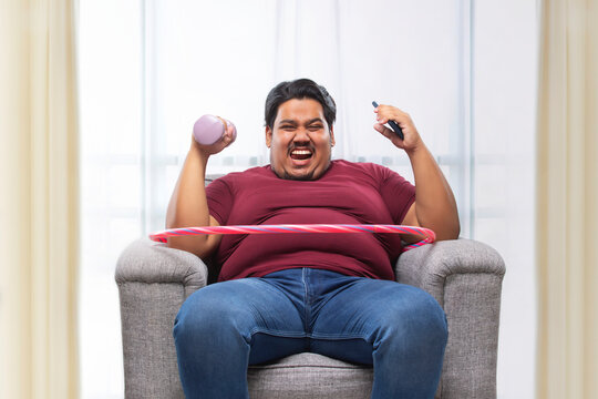 A fat man sitting on couch with dumbles and mobile with a hulla hoop around his waist.