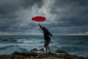 Young woman with a red umbrella on the background of the sea. Atmospheric autumn landscape with a young dark-haired girl in a developing dress. Storm, strong wind, overcast. The concept of happiness