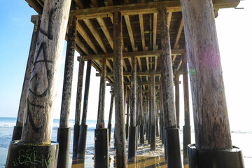 a shot of the legs underneath a brown wooden pier at the beach with waves rolling in from the vast...