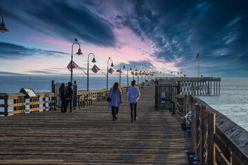 a long brown wooden pier with American flags flying on curved light posts with people walking and...