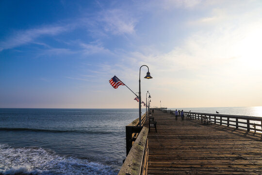 a long wooden pier with American flags and curved light posts along the pier with people walking along the pier and blue sky and clouds at sunset at Surfers Point at Seaside Park in Ventura California - Powered by Adobe