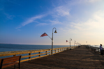 a long wooden pier with American flags and curved light posts along the pier with people walking along the pier and blue sky and clouds at sunset at Surfers Point at Seaside Park in Ventura California