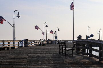 a long brown wooden pier with American flags flying from curved light posts and benches along the...
