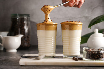 Male hand stirring trendy korean beverage dalgona coffee latte - whipped instant coffee, sugar and hot water and milk layers on a marble board, grey surface