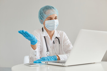 Indoor shot of helpless woman doctor wearing gown, surgical mask, medical cap and gloves, posing in front of notebook with spread aside hand, working in clinic.