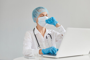 Fototapeta premium Unhealthy woman doctor therapist wearing gown, surgical mask, medical cap and gloves, sitting in front of computer in clinic and coughing, infected coronavirus from sick people.