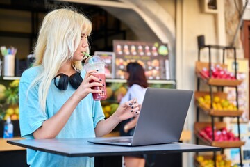 Teenage girl in fruit outdoor cafe drinking vitamin fresh juice, using laptop